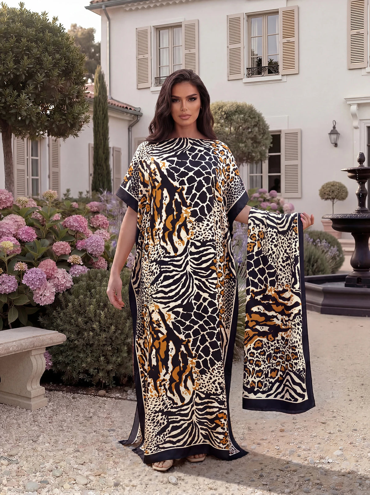 A woman wearing a black and gold leopard zebra print maxi kaftan standing outdoors in a garden setting.