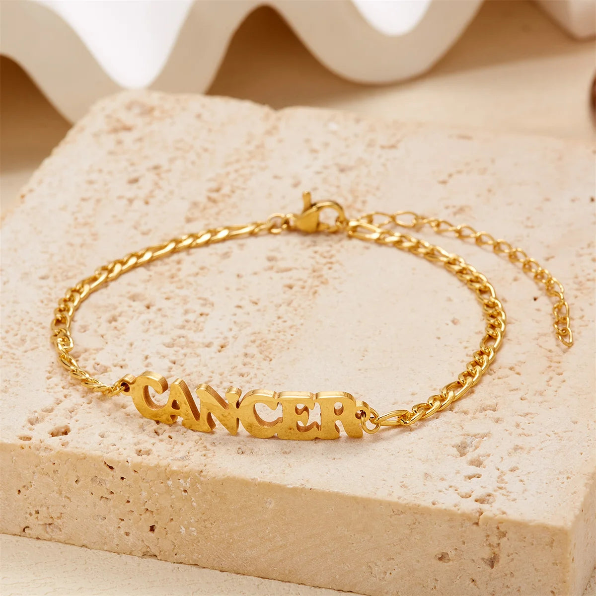 Gold stainless steel zodiac constellation charm bracelet spelling out CANCER resting on a light surface.