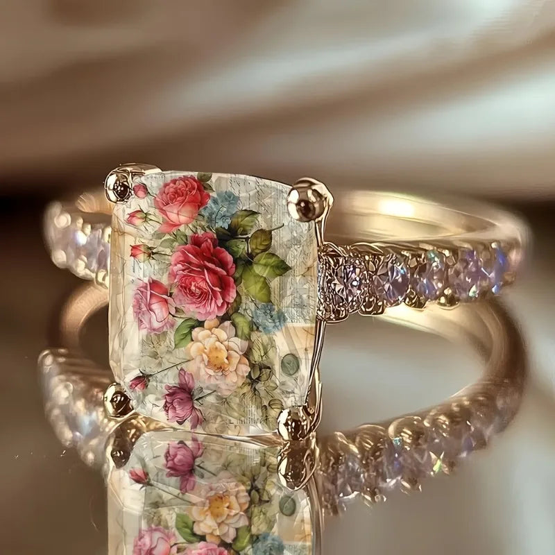 Bague argentée avec une pierre transparente carrée illustrant une rose rose sur une bande texturée.