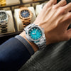 A hand wearing a silver-toned luxury quartz wristwatch with a blue luminous dial and stainless steel bracelet.