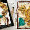 A silk scarf with a floral print featuring gold and teal leaves is displayed in a square frame.