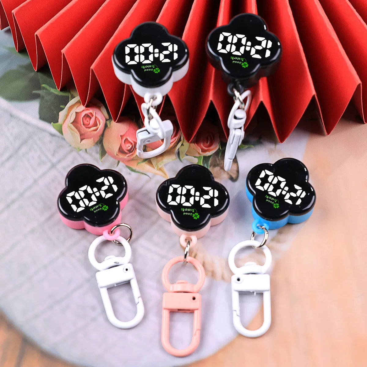 A set of black LED digital keychain watches with silver and pink charms displayed against a red pleated background.