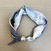 A light blue and white patterned silk scarf tied into a loop with a pearl tail magnetic clasp.