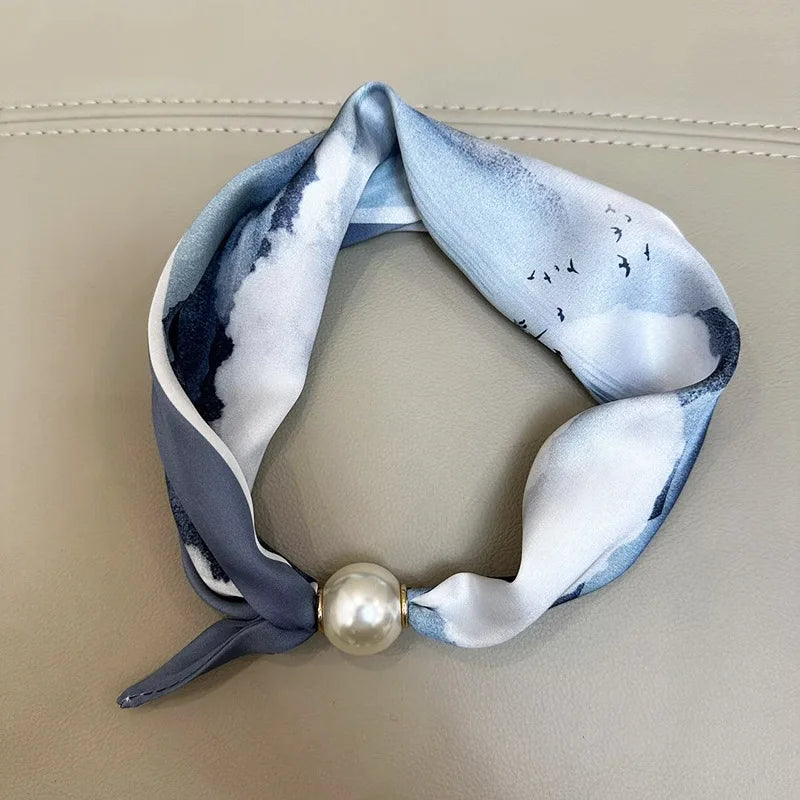 A light blue and white patterned silk scarf tied into a loop with a pearl tail magnetic clasp.
