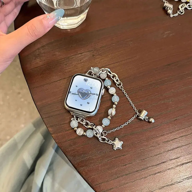 A silver double chain watch strap with clear beads and a heart-shaped charm resting on a wooden table.