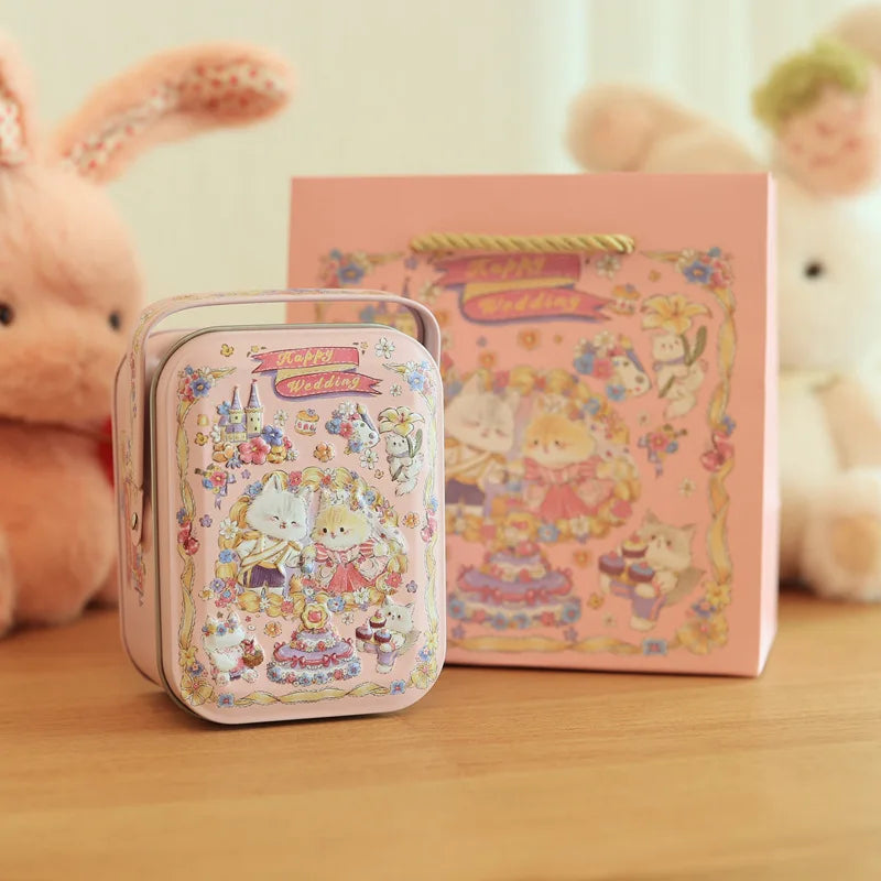 A pink decorative tinplate candy box with a carrying loop sits in front of a larger pink wedding-themed box and a fluffy rabb