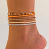 A stack of multi-layered beaded anklets in shades of orange, beige, and white worn on a person's ankle.