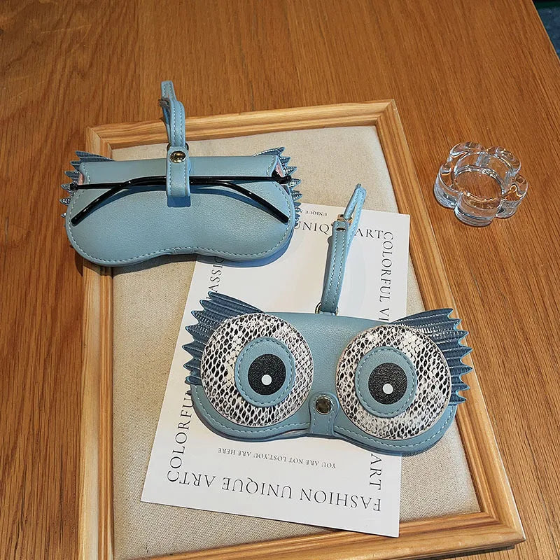 A light blue portable eyewear storage pouch with a decorative, textured frame is displayed in a wooden frame.