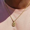 A gold-plated letter pendant necklace with the letter B worn around the neck against a pink shirt.