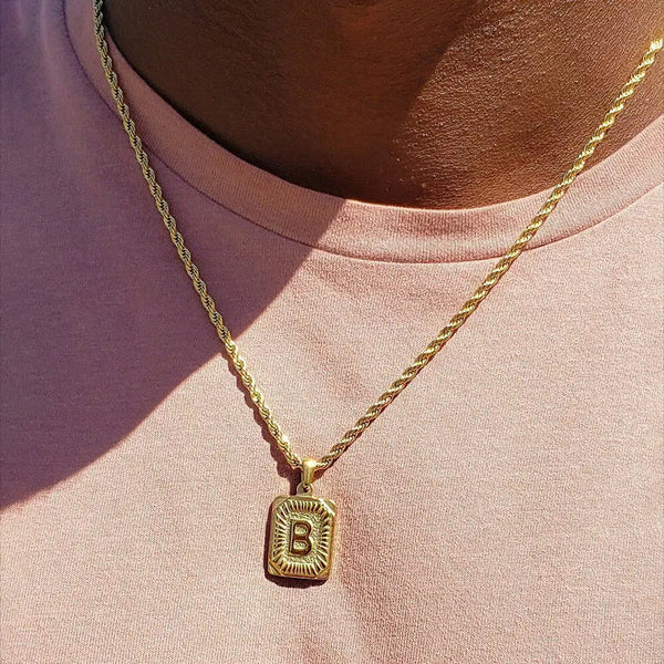 A gold-plated letter pendant necklace with the letter B worn around the neck against a pink shirt.