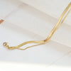 A delicate gold double snake anklet is shown against a white marble background.