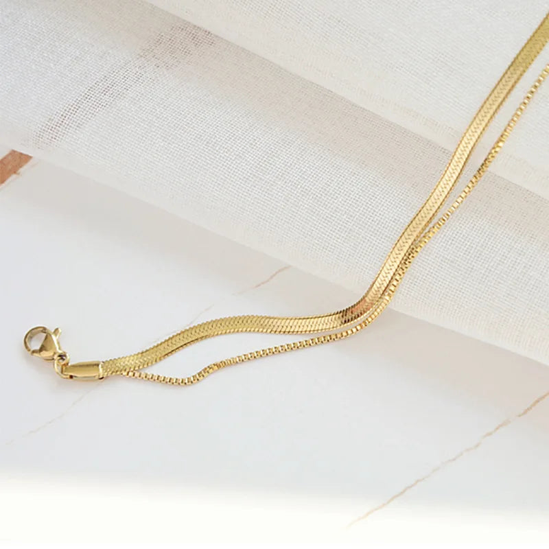 A delicate gold double snake anklet is shown against a white marble background.