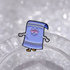 A small enamel cartoon brooch of a blue character with purple accents and surprised expression resting on a clear surface.