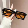 A hand holds a pair of black retro square sunglasses with amber lenses against a light background.
