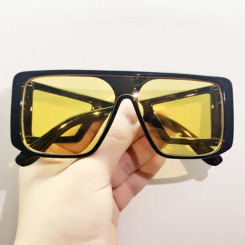 A hand holds a pair of oversized gradient square sunglasses with black frames and yellow-tinted lenses.