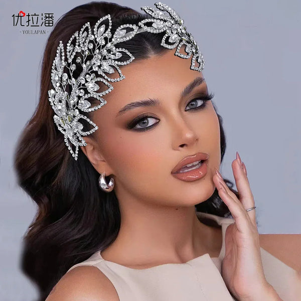 A close-up of a model wearing an intricate silver rhinestone leaf bridal hairband against a gray background.