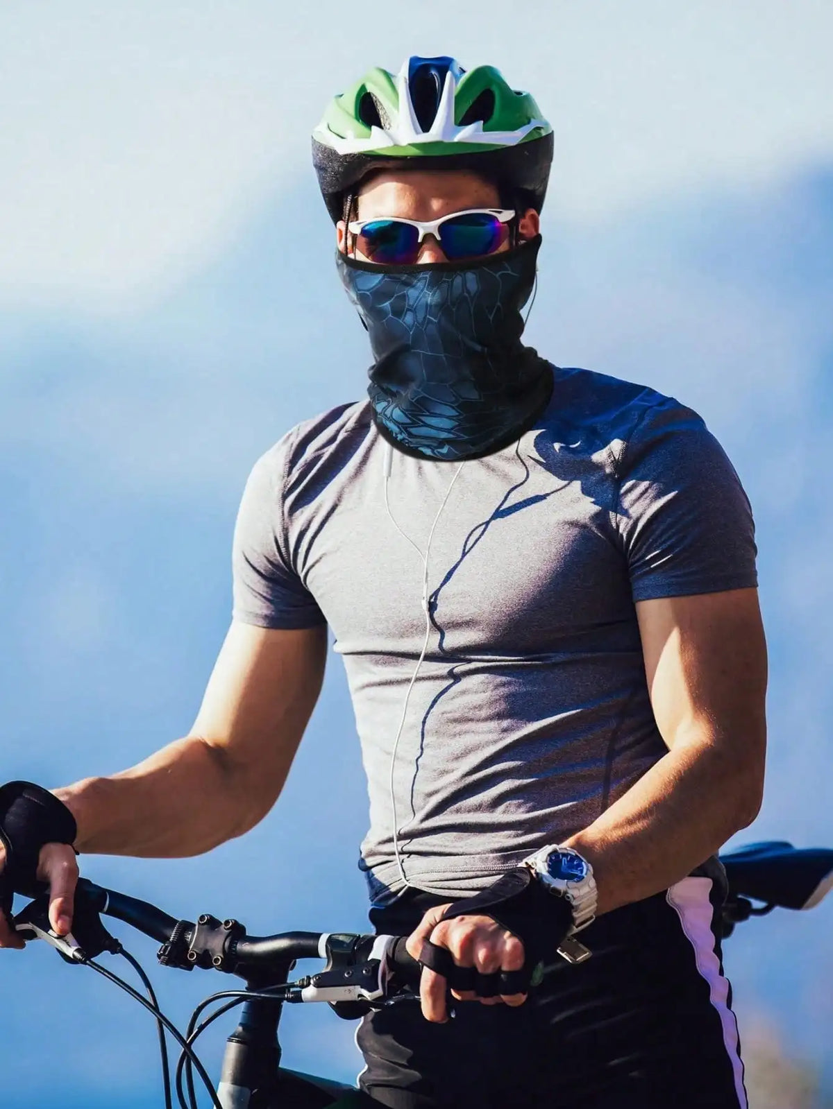 A man wearing sunglasses and a dark balaclava cap is riding a bicycle outdoors.