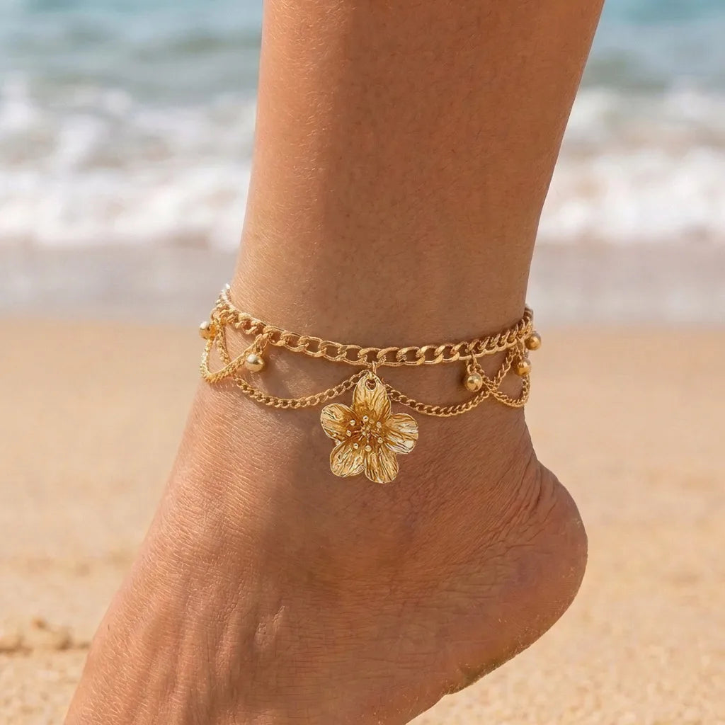 Gold chain anklet with a flower pendant worn on a person's ankle near the ocean.