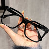 A hand holds a pair of black geometric reading glasses with transparent lenses against a light gray background.