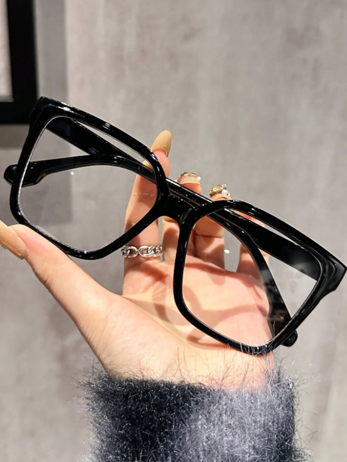 A hand holds a pair of black geometric reading glasses with transparent lenses against a light gray background.