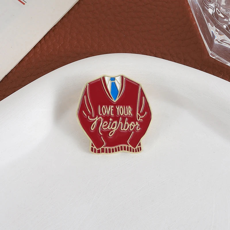 Broche émaillée rouge en forme de pull avec l'inscription 'Love Your Neighbor' en écriture blanche et un nœud papillon bleu.