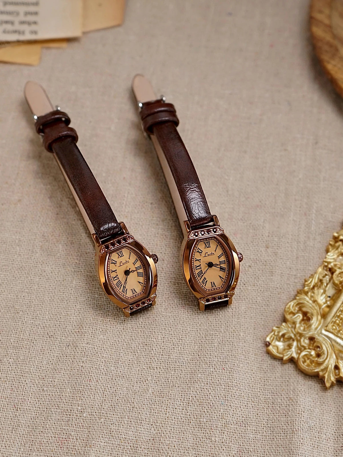 Un par de relojes de cinturón de cuero marrón retro con cajas doradas y decoración de strass sobre tela beige.