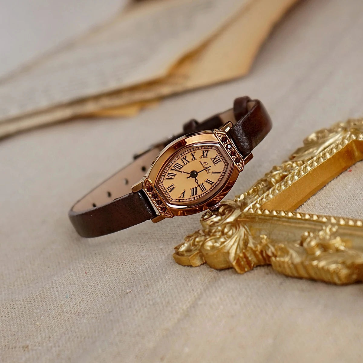Reloj de correa de cuero marrón retro con detalles dorados y pedrería, sobre tela clara.
