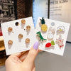 A hand holds two white metal pin badges featuring cute animal and plant designs like a pineapple, rabbit, and dinosaur.
