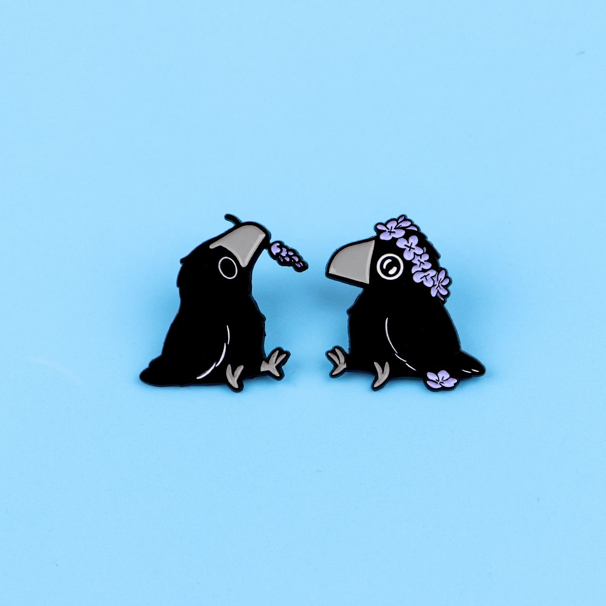 Ein Paar schwarze Email-Anstecknadeln mit Raben und lila Blumen.