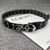 Black studded leather belt with a silver skull buckle displayed on a white card.