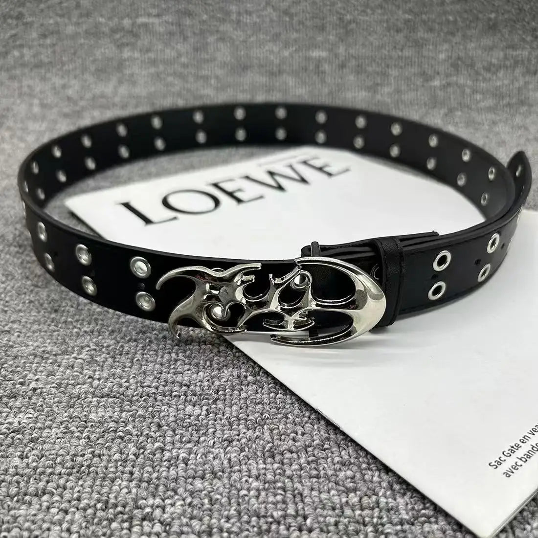 Black studded leather belt with a silver skull buckle displayed on a white card.