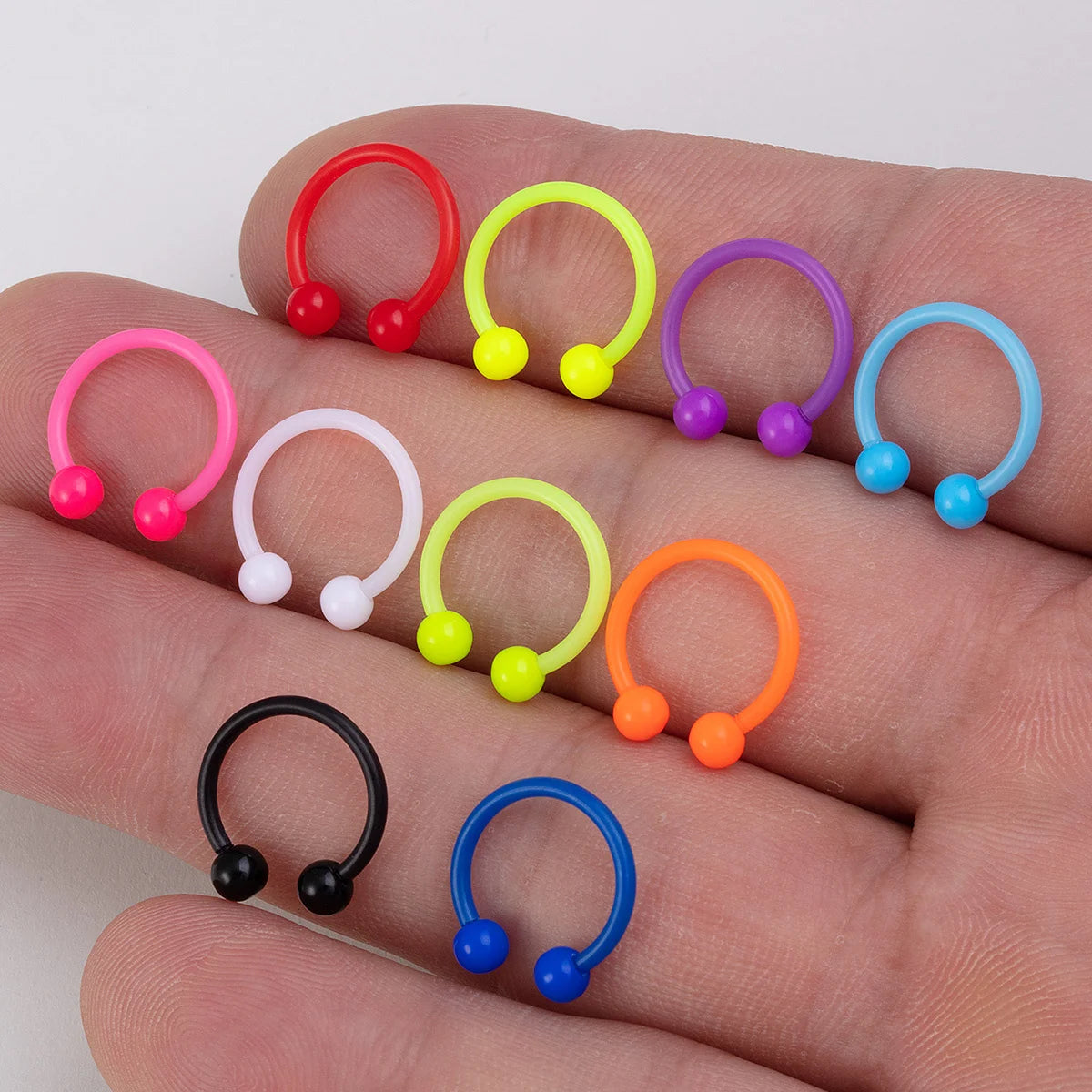 A hand displays a variety of colorful flexible acrylic piercing jewelry rings and posts.