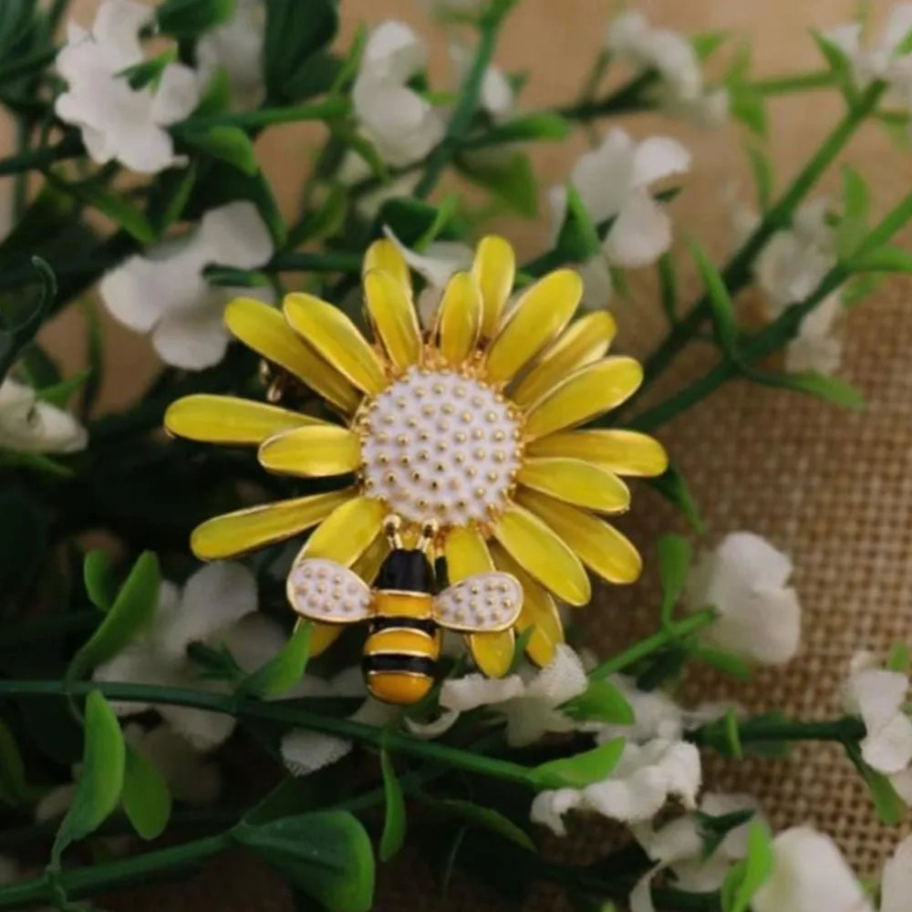 Broche florale dorée en forme d'abeille avec détails blanc et noir, sur fond de feuillage vert.