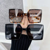 A hand holds a pair of brown leopard print square sunglasses and a pair of black square sunglasses.