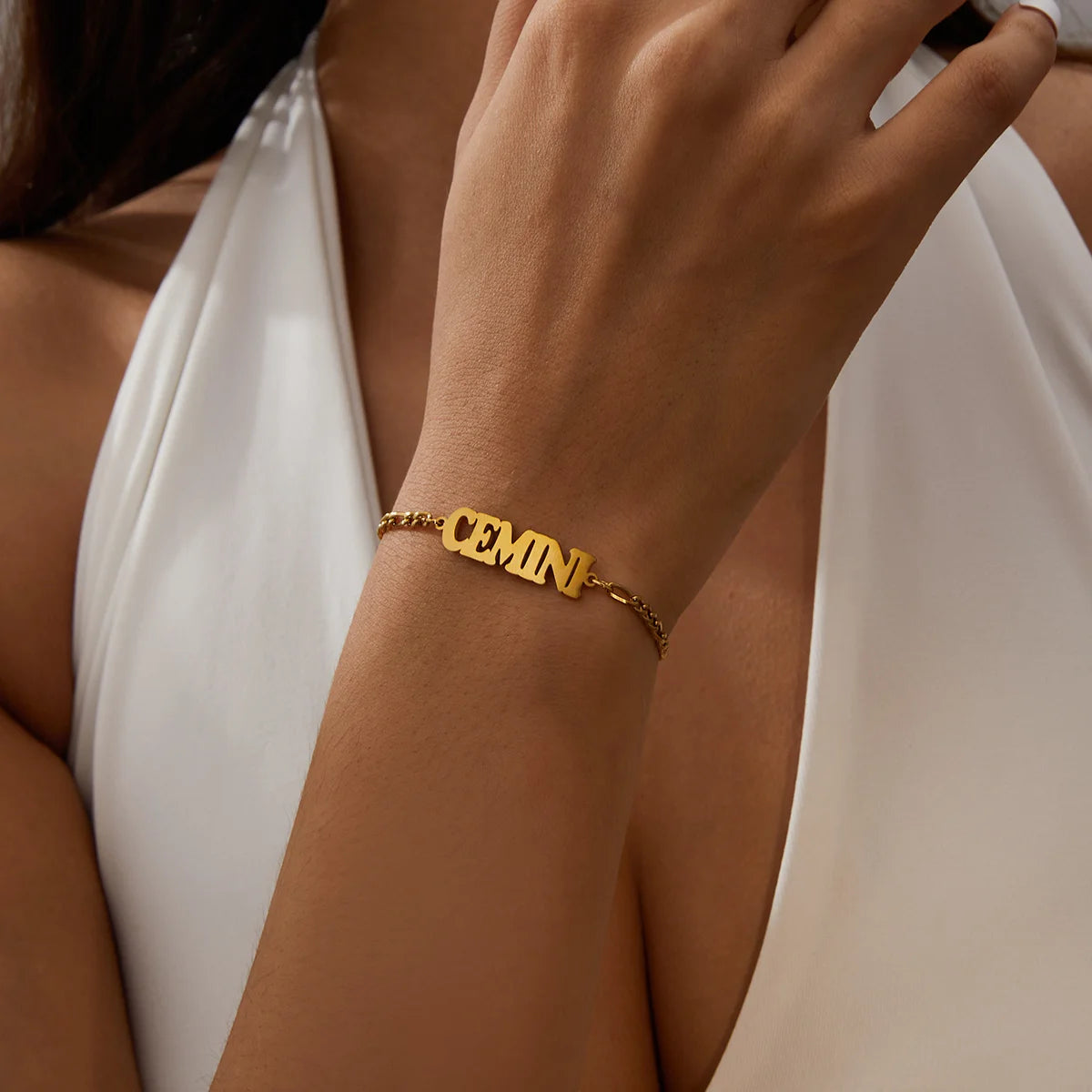 A close-up of a hand wearing a gold stainless steel Zodiac constellation charm bracelet on a white dress.