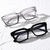 A pair of clear, square-framed reading glasses resting on a white textured surface.