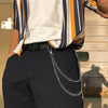 Una persona vistiendo una camisa blanca a rayas verticales y pantalones oscuros, con una cadena metálica de cintura.