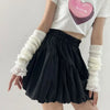 A person wearing black pleated skirt and white fingerless arm warmers with a pink heart graphic on their white top.