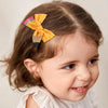A young girl with brown hair wears a yellow and pink pencil bow hair clip on the side of her head.