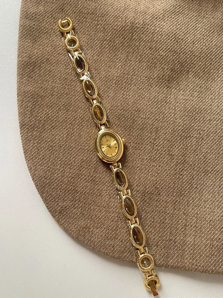 Montre de quartz dorée, ronde, avec bracelet en maillons, présentée sur un fond de tissu marron.