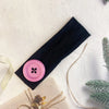 A black hairband with a pink button detail is displayed on a light background next to natural twine and greenery.
