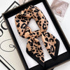 A black and tan leopard print silk scarf tied in a knot, displayed in a black frame.