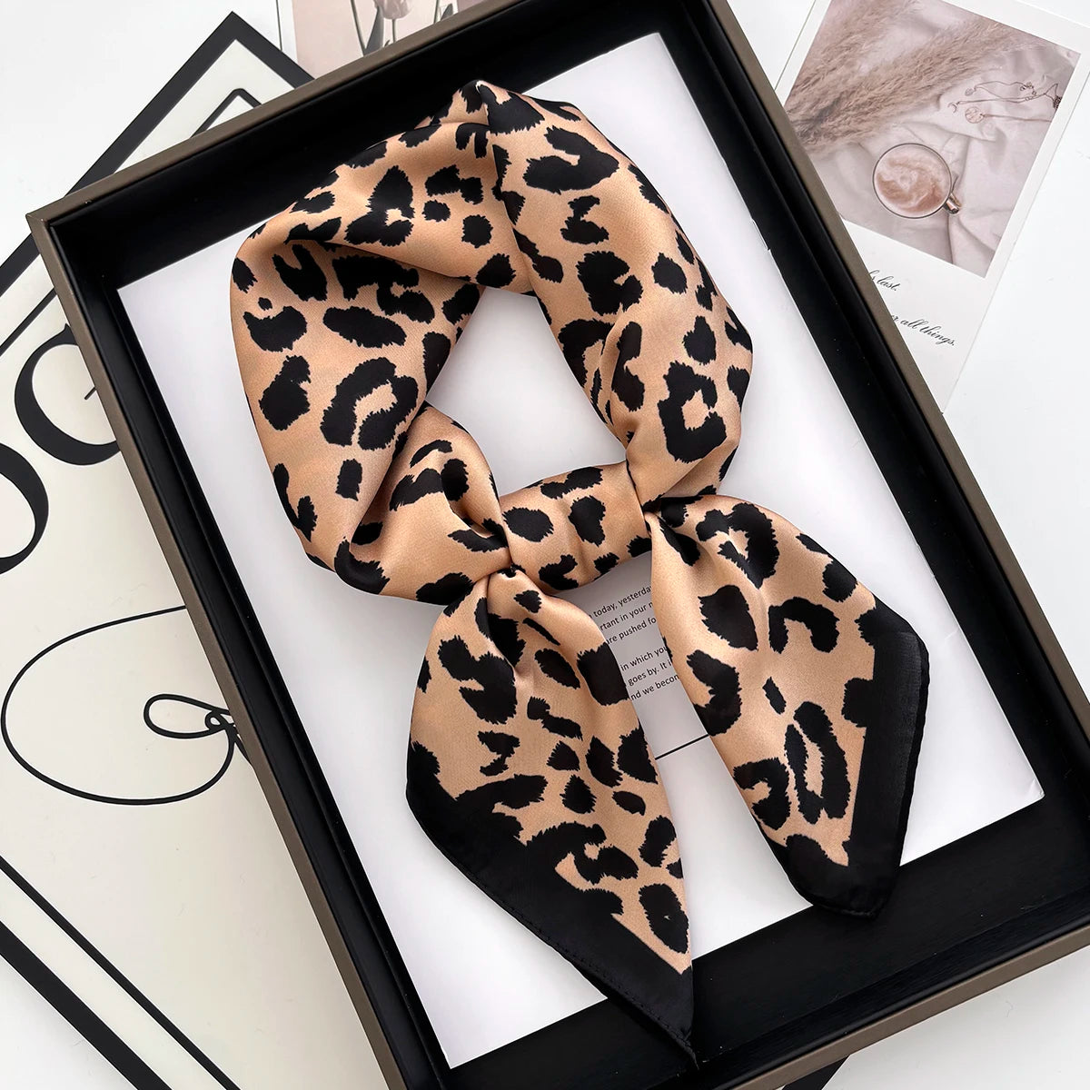 A black and tan leopard print silk scarf tied in a knot, displayed in a black frame.