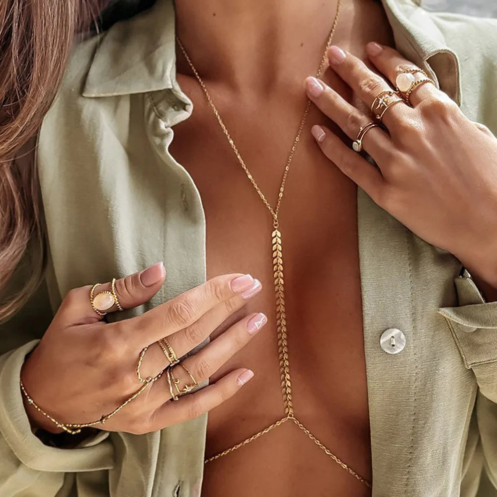 A close-up of a woman wearing a gold minimalist wheat body chain necklace and rings, paired with an olive green shirt.