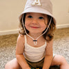 A smiling toddler wearing a beige letter print sun hat and a white tank top sitting on a carpet.