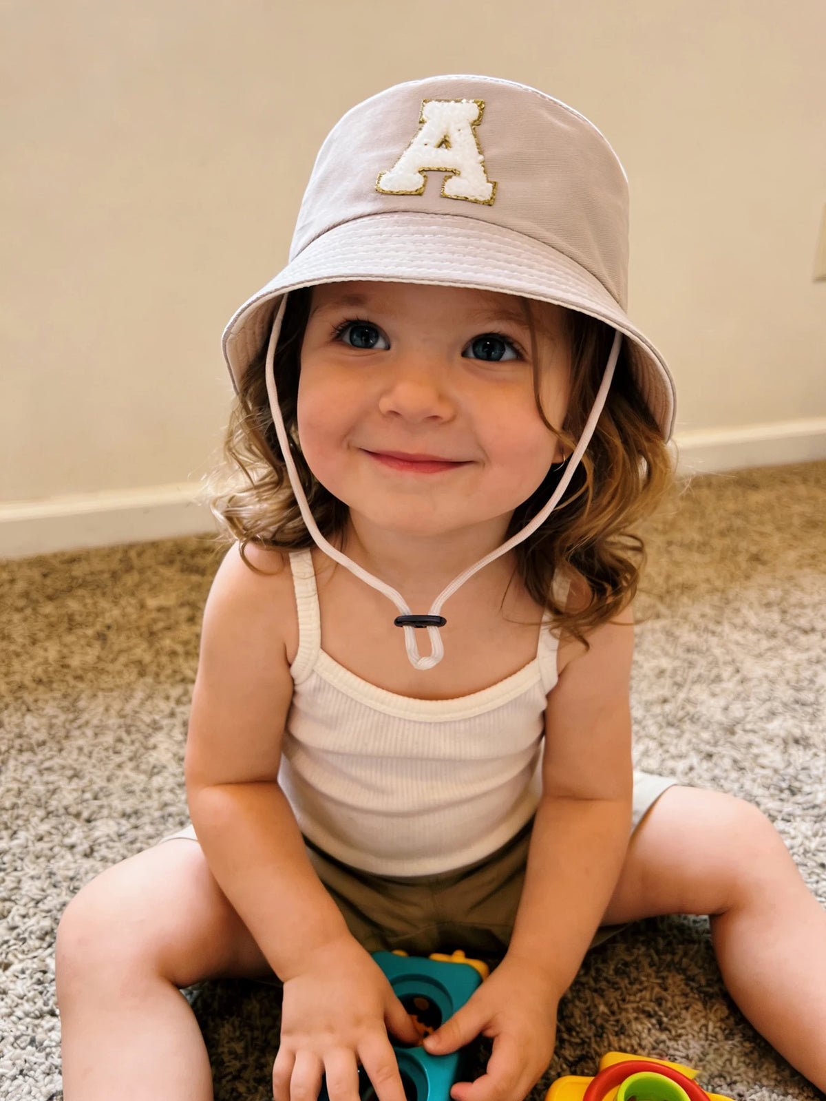 A smiling toddler wearing a beige letter print sun hat and a white tank top sitting on a carpet.