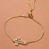 A delicate gold plated cross bracelet with cubic zirconia stones on a light pink background.