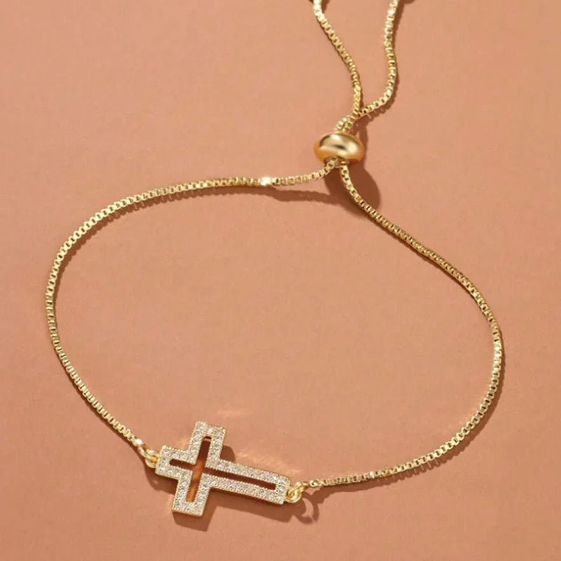 A delicate gold plated cross bracelet with cubic zirconia stones on a light pink background.