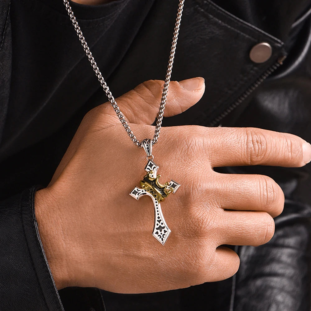 A hand wearing a silver necklace with a gold crown cross pendant against a black leather jacket.