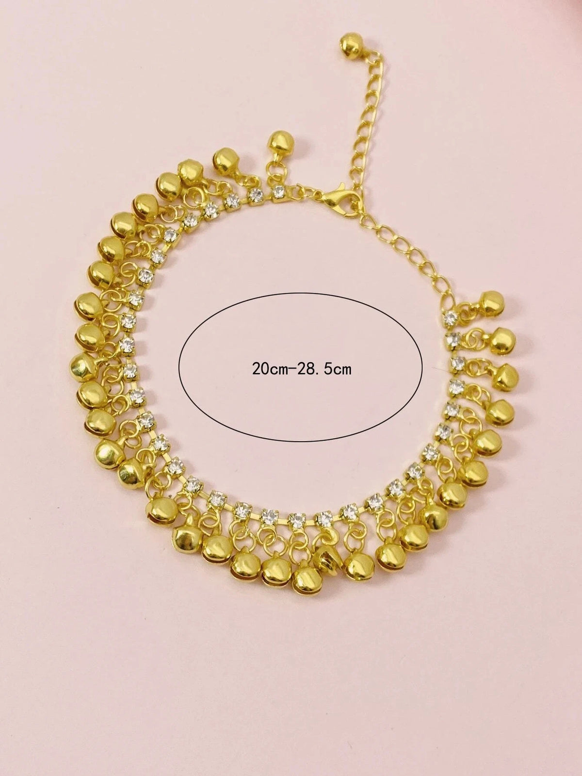 A gold bell foot chain anklet with small clear stones, shown against a light pink background.
